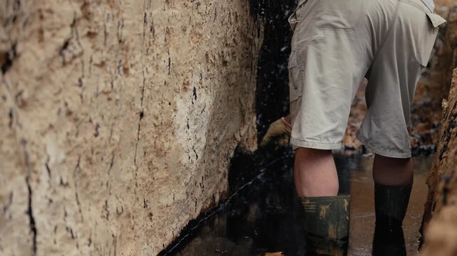 Male worker standing in a trench and using a brush to apply black tar waterproofing sealant to the exterior of a building foundation, ensuring damp proofing and structural integrity