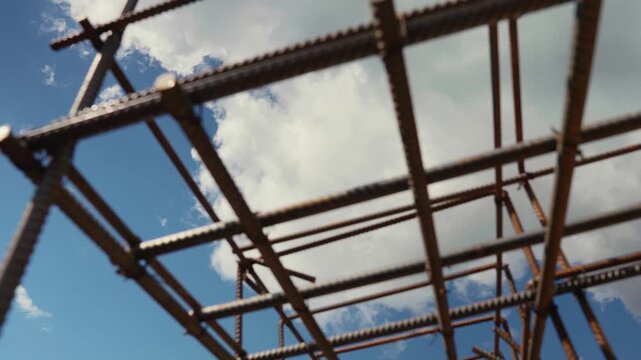 Low angle perspective of a rusty steel rebar cage for concrete reinforcement on a construction site, with a beautiful blue sky and white cumulus clouds moving in the background