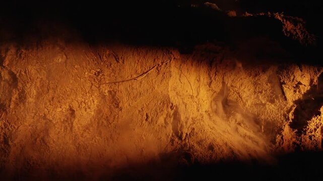 Billowing dust and debris erupting from a warm, orange lit earthen wall during a collapse. The dramatic, slow motion footage captures the texture and particles of the soil as they explode