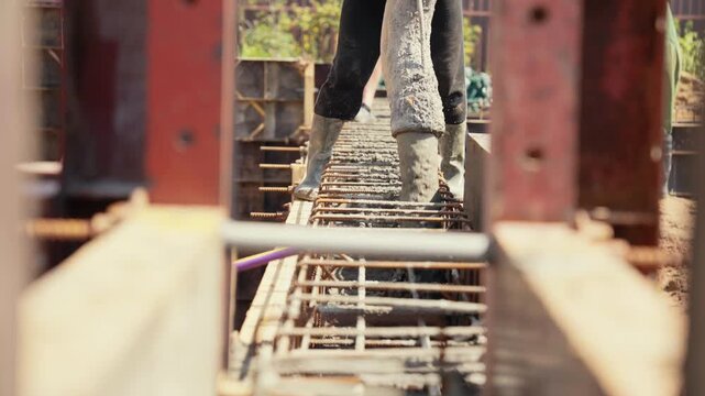 Concrete pouring from a large hose into a steel reinforcement cage on a construction site, with a worker in rubber boots supervising the process of laying the foundation for a new building
