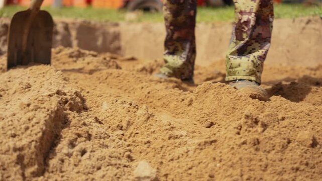 Military soldier wearing camouflage uniform digging a trench with a shovel during a training exercise. The serviceman is moving sand, fortifying a defensive position in a wartime simulation