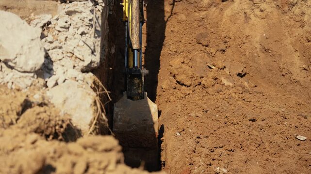 Mechanical excavator digging a deep trench in the soil at a construction site for laying pipes or foundations, with the bucket moving earth and rubble during groundwork operations on a sunny day