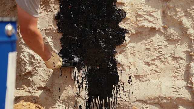 Male builder hand in a protective glove using a brush to apply a thick black bitumen sealant for waterproofing a concrete foundation wall at a construction site in slow motion