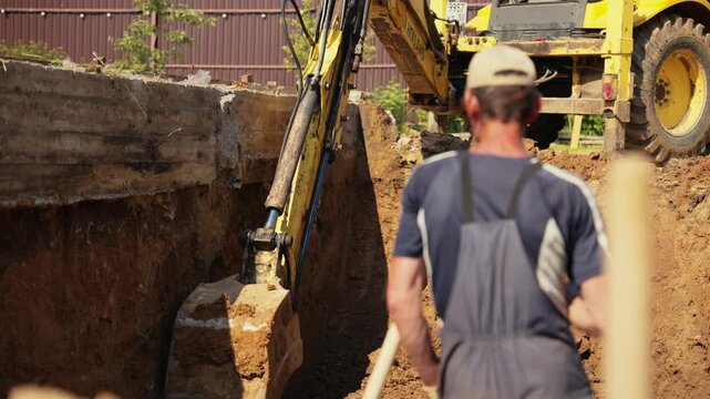 Heavy machinery excavating a deep pit for a building's foundation on a construction site, while a worker in overalls stands by and supervises the earthmoving operation during a sunny day