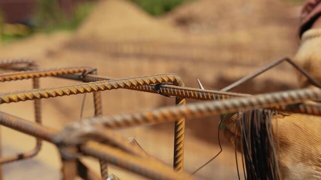 Close up view of a builder's gloved hands using a special hook tool to tie together rusty steel reinforcing bars with thin wire, preparing the framework for pouring a concrete foundation