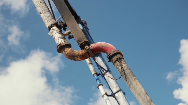 Detail of a mobile concrete pump truck boom arm extending and retracting with its hoses and rusty joints, viewed from below against a bright blue sky with scattered white clouds on a sunny day