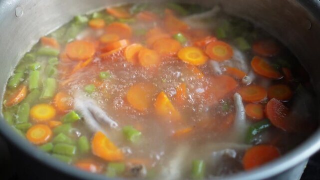 Boiling chicken feet soup with sliced carrots and green beans in a pot steam rising from bubbling broth rich with vegetables and meat for healthy cooking or restaurant use.