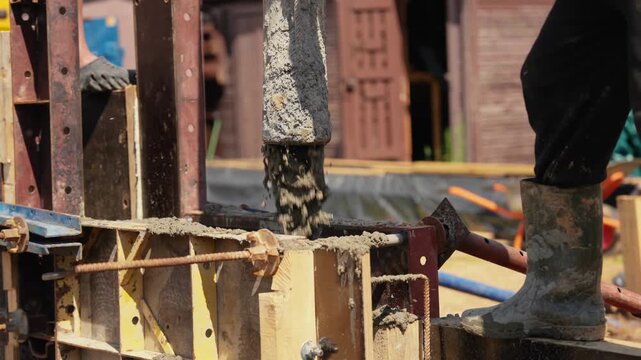 Wet cement pouring from a pump hose into a wooden formwork structure on a building site, with a construction worker in rubber boots standing nearby during the foundation casting process