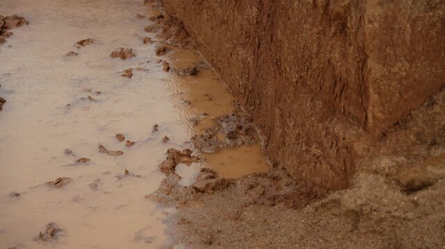 Heavy rain falling on a dirty puddle during a downpour, creating ripples in the murky brown water as it accumulates against the earthy soil and wet sand on a stormy and wet day