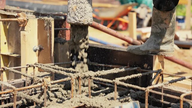 Liquid concrete being poured from a pump hose into a wooden formwork with a metal rebar cage, creating a reinforced foundation for a new building during construction work on a sunny day