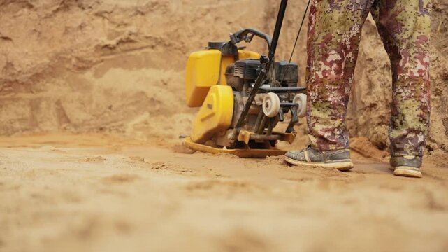 Closeup of a construction worker in camouflage pants operating a vibratory plate compactor, leveling and tamping sand at a building site during groundwork and foundation preparation