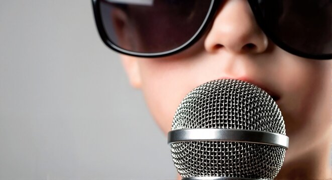 Close up of caucasian child wearing sunglasses singing into toy microphone. Kid performing with silver mic on gray background. Copy space