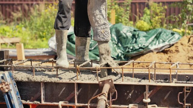 Professional builder in rubber boots pouring liquid cement mix from a pump hose into a metal formwork with steel rebar to create a solid reinforced concrete foundation for a new house