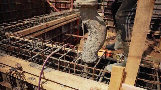 Slow motion view of a construction worker using a concrete pump to pour wet cement mixture into a reinforced steel cage, creating the foundation for a new residential building