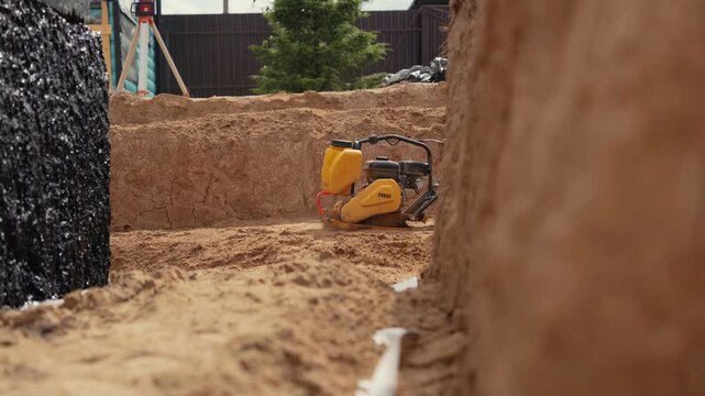 Industrial equipment tamping and leveling sand at the bottom of a foundation trench. Close up view of a vibratory plate compactor working on soil in slow motion during building preparation