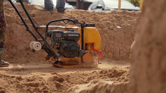 Construction worker using a vibratory plate compactor for soil compaction at a building site, tamping sand to prepare the foundation for paving and raising clouds of dust during the process