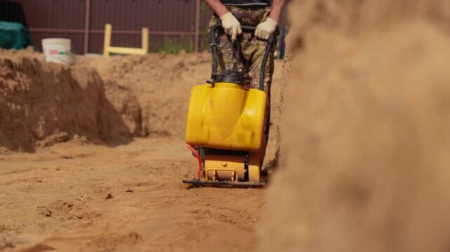 Male worker using a tamping machine for soil compacting at a construction site before laying the foundation. Ground preparation process with a yellow professional vibratory plate compactor