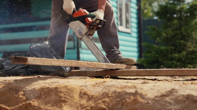 Construction worker wearing protective gloves using a powerful chainsaw to cut through a wooden plank at an outdoor worksite, with sawdust flying in the air during the process