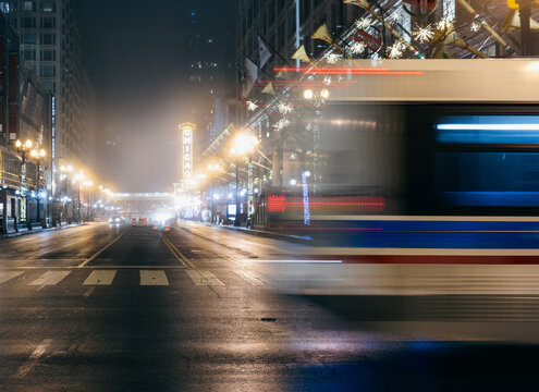 A bus rushes through a lively street in Chicago, illuminated by bright lights and holiday decorations, capturing the essence of urban mobility on a foggy evening.
