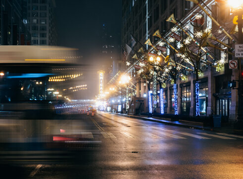A calm, foggy street scene in Chicago comes alive with festive holiday lights, creating a serene yet lively atmosphere amidst the blurred motion of passing vehicles.