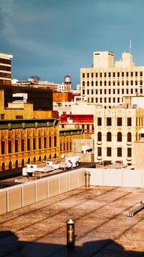 Urban buildings stand close together in a busy area. The sun sets in the background, casting light on the rooftops and walls. Construction activities can be seen on some roofs.