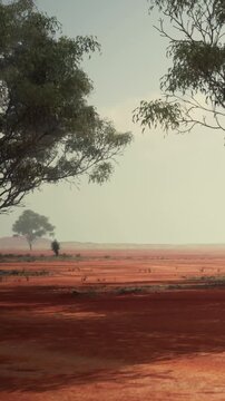 A landscape shows red earth with several trees scattered in the view. The sky is light and slightly cloudy. The scene is wide and open, showing a natural setting.