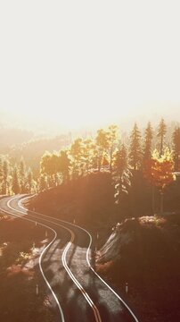 A road curves through a landscape filled with trees as the sun sets in the background. The warm light highlights the natural beauty of the surroundings in autumn.
