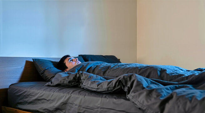 Young man lying awake in bed with insomnia. Person staring at ceiling under dark gray sheets in dimly lit bedroom