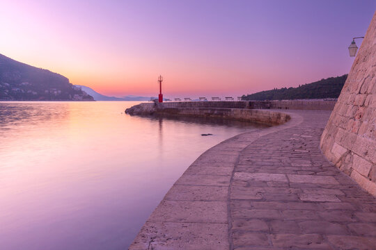 Curved stone waterfront promenade leading to a harbor lighthouse at sunrise in Dubrovnik, Croatia, with calm Adriatic Sea and soft morning light.