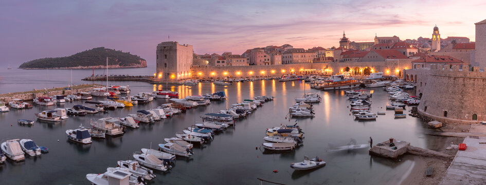 Panoramic view of Dubrovnik Old Harbor with boats, medieval city walls, and illuminated historic buildings at blue hour in Dubrovnik, Croatia.