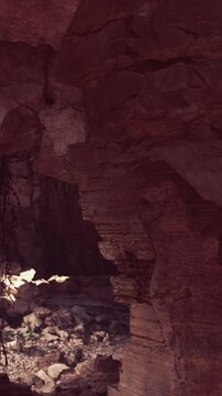 Sunlight enters a canyon through rock formations, creating shadows on the ground. Plants grow near the rocks. The scene shows natural textures and colors typical of a dry area.