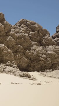 Large rocks are present in a desert landscape. The ground is sandy, and there is a bright blue sky above. The scene shows natural formations and dry terrain during daylight.