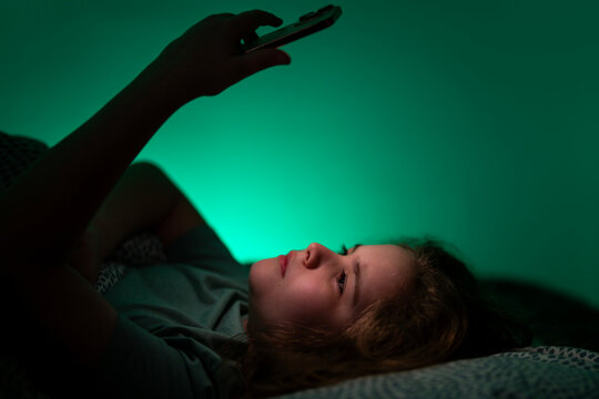 Child lying comfortably while using smartphone. Kid browsing online content in bedroom. Child experiencing modern digital bedtime habit. Kid using phone quietly in dark room.