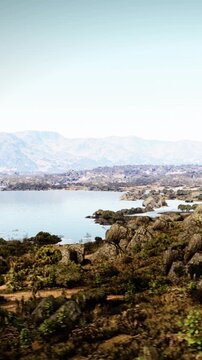 A clear view of a lake surrounded by rocky land and distant mountains. The water reflects the blue sky. Vegetation is visible near the shoreline under bright sunlight.