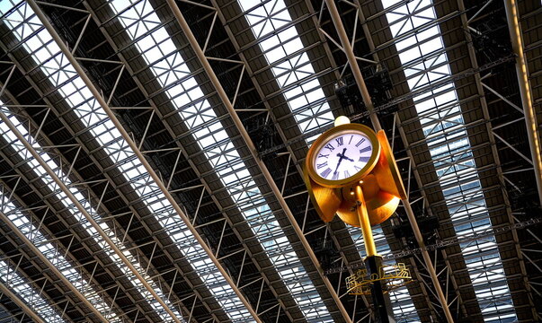 大阪駅の広大なドーム屋根の下に設置された金色の巨大な塔時計。モダンな鉄骨構造と幾何学的なガラス天井が特徴的な駅構内の風景。