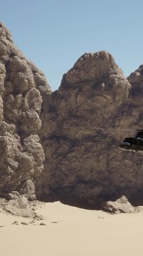 A helicopter hovers near large rocky formations in a dry desert. The sun is high in the clear blue sky, casting shadows on the sandy ground below.