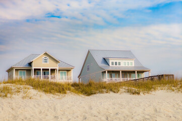 Two beach houses nestled in dunes under a cloudy blue sky © EJ