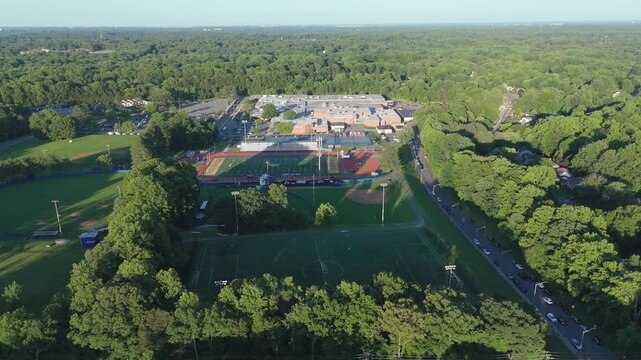 Aerial drone shot of a large suburban high school campus featuring a football stadium, baseball diamonds, and soccer fields surrounded by lush green summer trees and a local road.