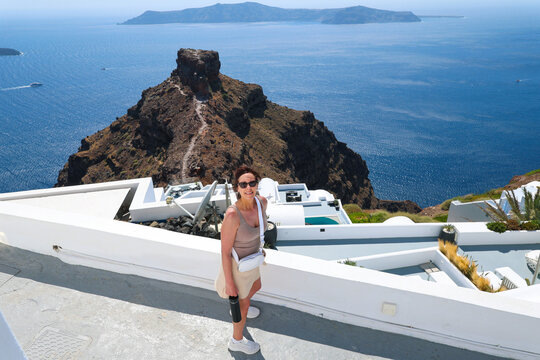 A woman stands on a white terrace overlooking Skaros Rock and the deep blue Aegean Sea in Santorini, Greece, surrounded by bright sunlight and stunning coastal views.