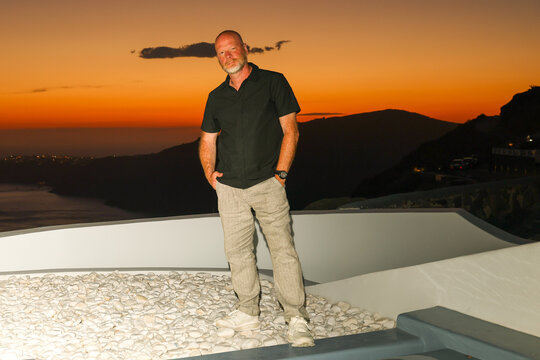 A bald man stands on a white terrace under a vivid orange sunset sky in Santorini, Greece, capturing the calm and color of Mediterranean evening light.