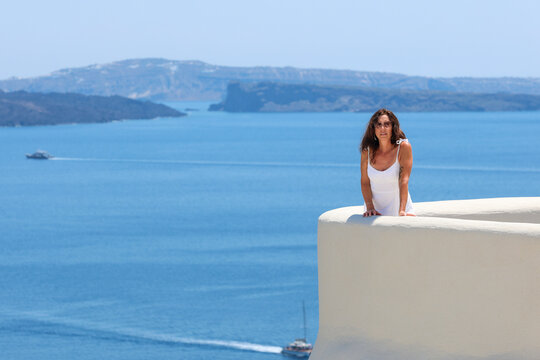 A woman in white stands on a curved terrace overlooking the Aegean Sea in Santorini, Greece, surrounded by bright sunlight and serene coastal views.