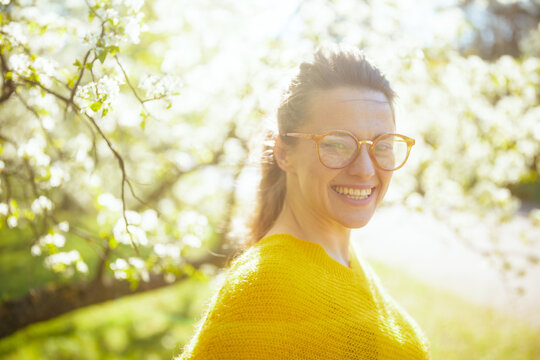 Looking directly at the camera with a wide smile, a woman in a yellow sweater and glasses enjoys a sunlit day surrounded by blooming spring trees.