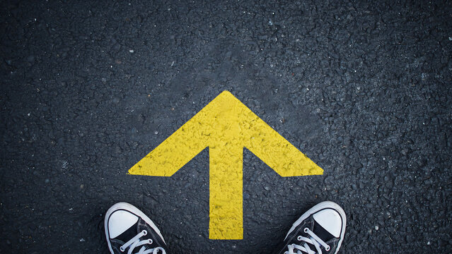 Person standing on asphalt road facing yellow painted upward arrow with direction decision movement lifestyle sneakers navigation motivation standing adventure