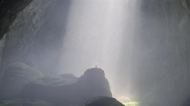 Closeup shot of a person standing a big stalagmite with a giant column of light at the background