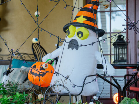 A large inflatable Halloween ghost wearing a striped witch hat holds a glowing pumpkin bucket on a decorated porch. Spider webs, hanging eyeballs, spiders, and purple lights create a festive Halloween