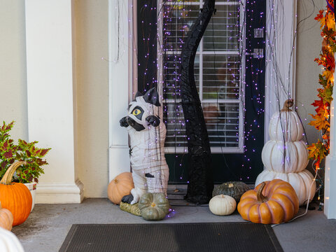 Pumpkins, decorative lights, and Halloween ornaments decorate a welcoming residential front entrance. A mummy cat figure and stacked pumpkins create a festive autumn holiday scene.