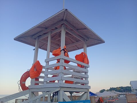 Wooden lifeguard tower painted white on a public beach, equipped with bright orange life jackets and a lifebuoy against a clear blue evening sky.