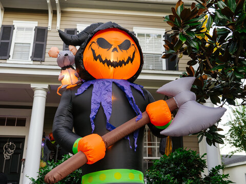 A giant inflatable Halloween reaper with a glowing pumpkin head and large axe stands in front of a residential house. The festive outdoor decoration creates a spooky Halloween atmosphere for the
