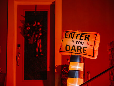 A glowing Halloween warning sign reading "Enter If You Dare" stands outside a decorated residential entrance at night. Orange lighting creates a spooky atmosphere for Halloween celebrations.