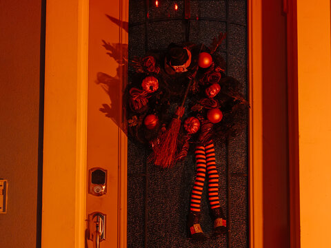 A Halloween wreath with pumpkins, a witch hat, and striped legs hangs on a front door illuminated by orange light. The festive decoration creates a spooky nighttime entrance.